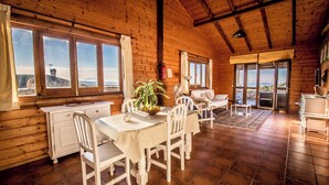 Una cabaña de madera con comedor, muebles blancos y ventanas amplias con vistas al océano.