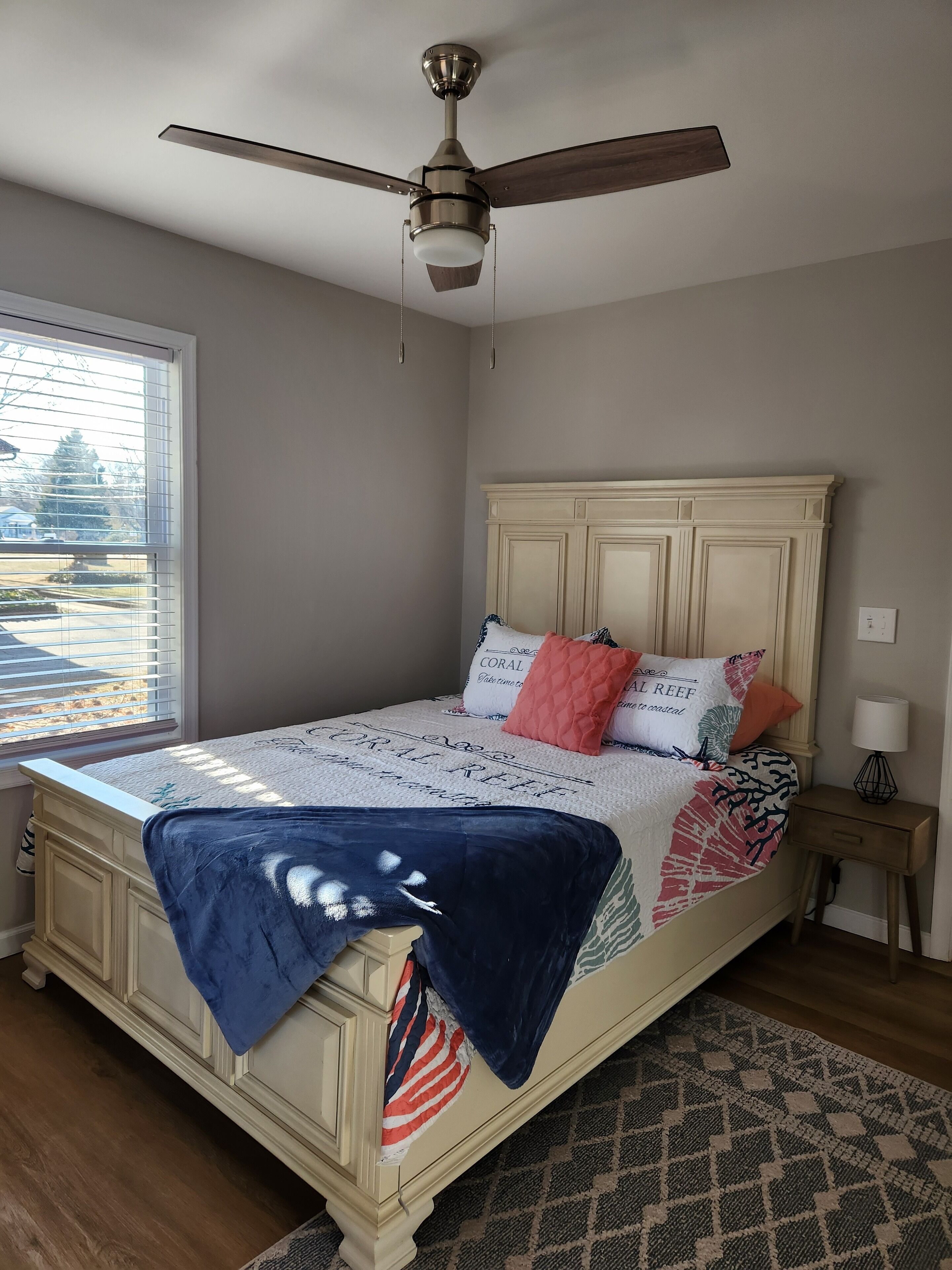Quarto com cama, ventilador de teto, janela com persianas, mesa de cabeceira com abajur e tapete estampado.