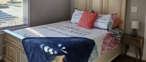 Quarto com cama, ventilador de teto, janela com persianas, mesa de cabeceira com abajur e tapete estampado.