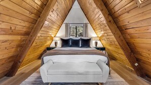 An attic bedroom with a sloped ceiling, a bed with a white headboard, two bedside lamps, and a window with curtains. - Kachina A-Frame Hideaway | AC | Hot Tub | Firepit (Flagstaff)