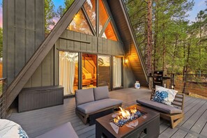 Een modern houten A-frame hutje met een terras, buitenmeubels en een vuurplaats.