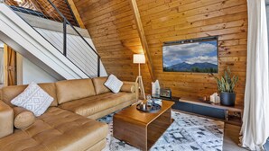A cozy loft bedroom with a sloped ceiling, a wooden staircase, a brown leather sofa, a wooden coffee table, and a large window with a mountain view. - Kachina A-Frame Hideaway | AC | Hot Tub | Firepit (Flagstaff)