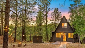 A cabin with a triangular roof, surrounded by trees, and a pathway leading to the entrance. - Kachina A-Frame Hideaway | AC | Hot Tub | Firepit (Flagstaff)