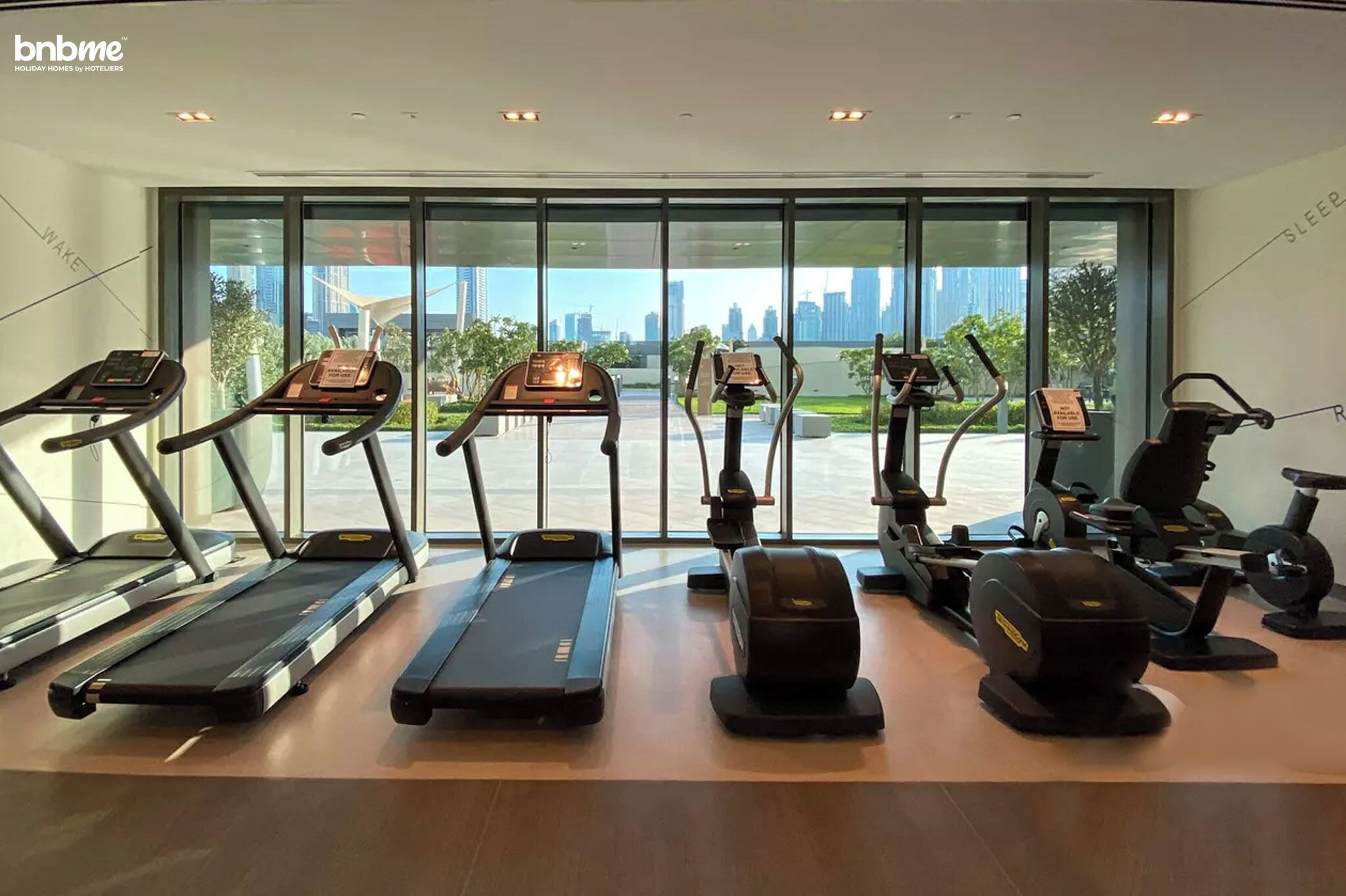 A gym with treadmills, elliptical machines, and a stationary bike, with a city view through large windows.