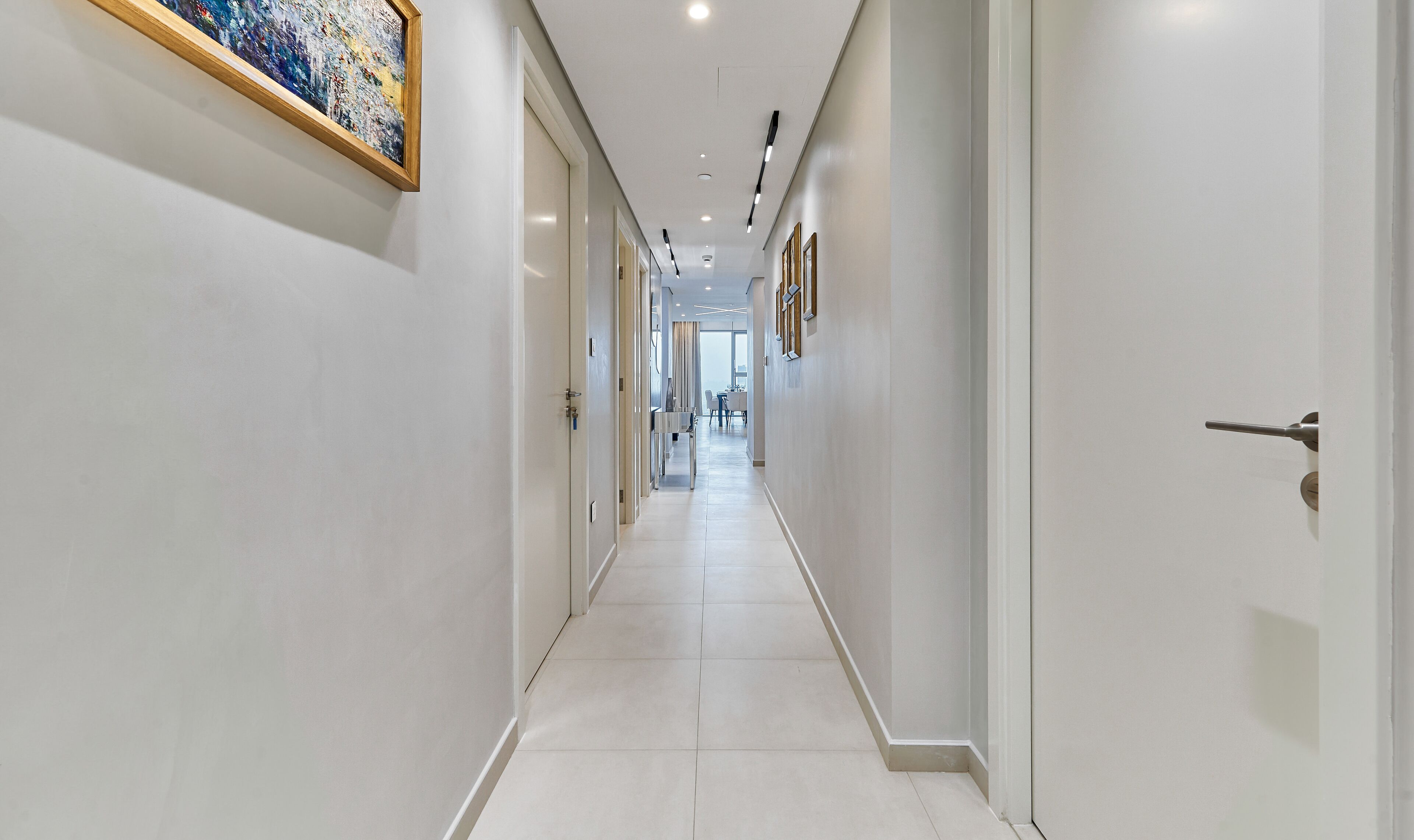 A modern hotel corridor with a long, narrow layout, recessed lighting, and a dining area visible in the distance.