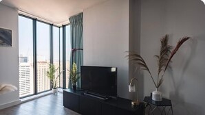Una sala de estar moderna con un ventanal grande, un televisor de pantalla plana sobre un mueble negro y una mesita auxiliar con una lámpara y plantas decorativas.