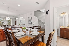 A dining area with a wooden table set for four, surrounded by chairs. There is a kitchen area with a countertop and cabinets visible in the background.
