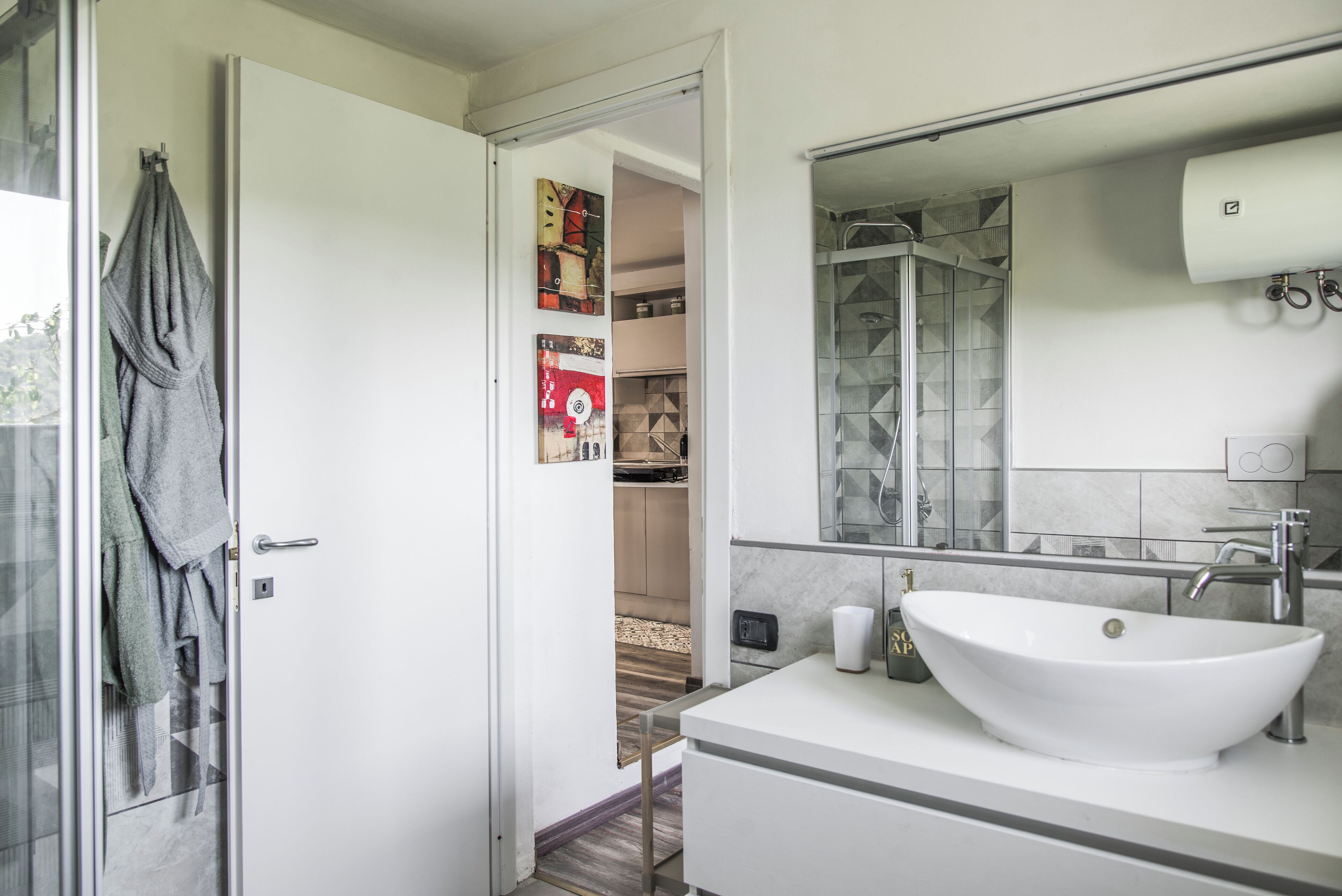 A modern bathroom with a large white sink, a glass shower enclosure, and a visible hallway leading to a kitchen area.