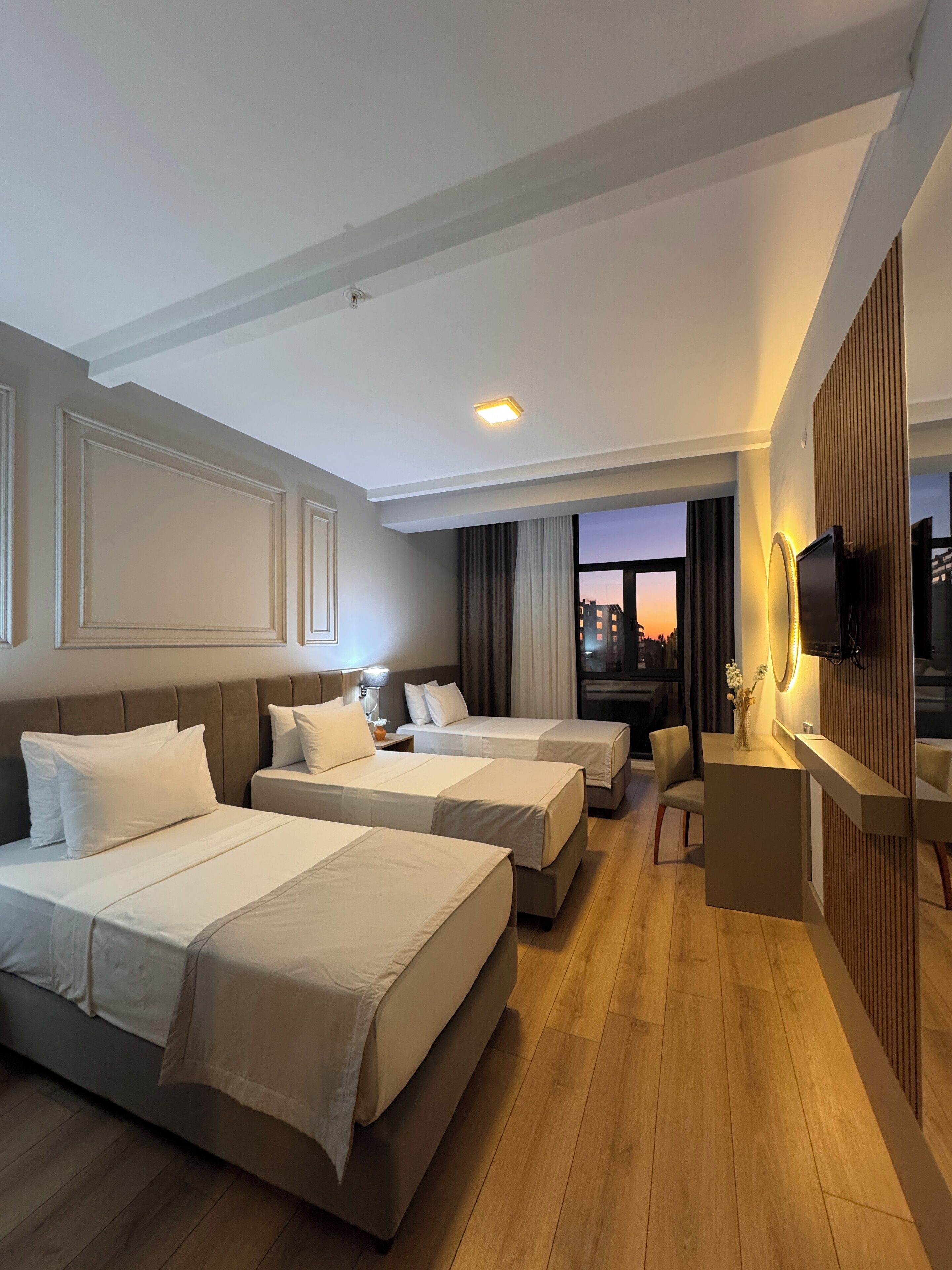 A hotel room with two beds, a desk, a TV, and a window with a city view.