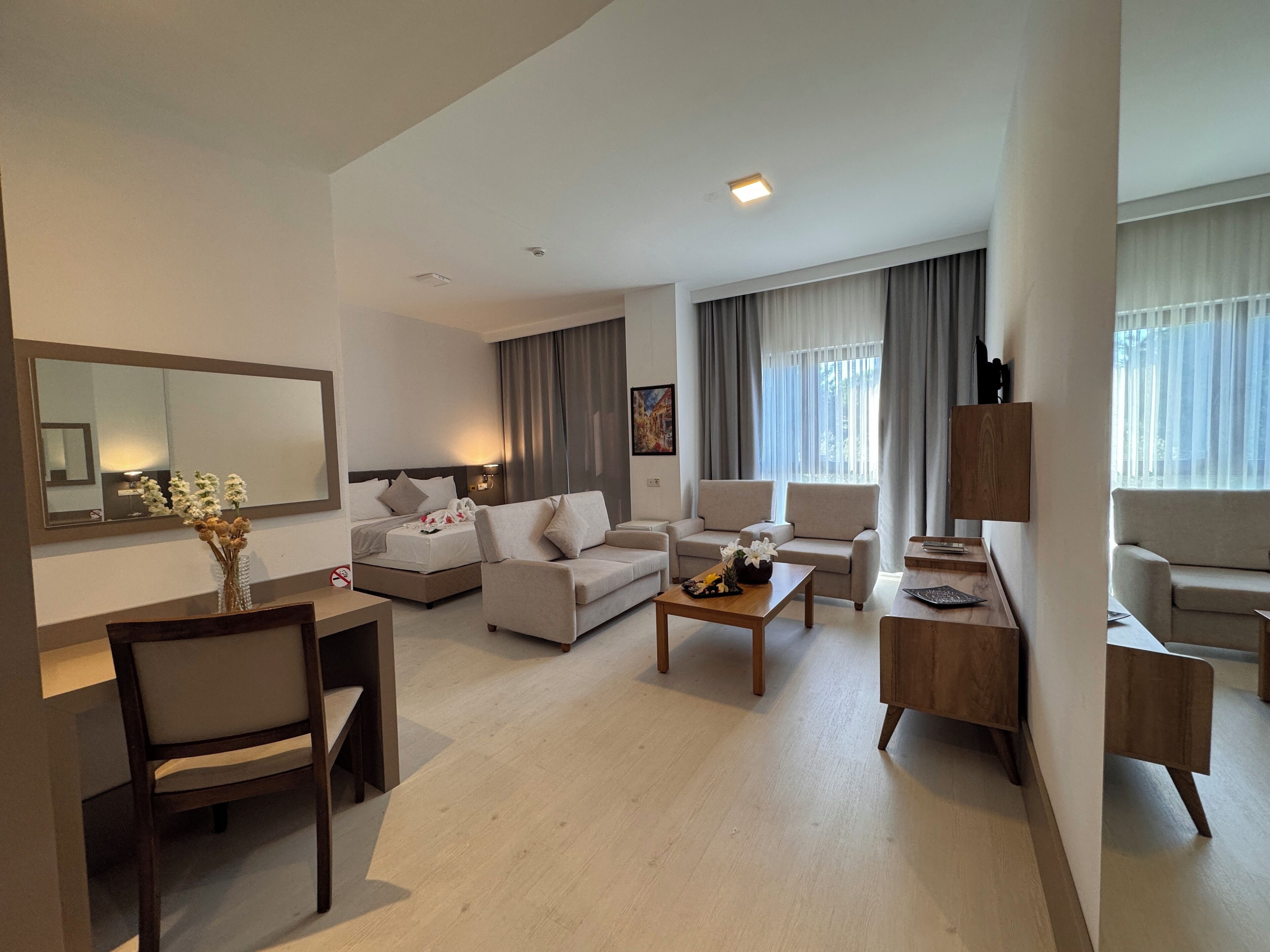 Una habitación de hotel moderna con una cama, dos sofás, una mesa de centro, una mesa de comedor con sillas y un amplio ventanal con cortinas.