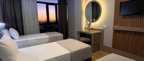 Habitación de hotel con dos camas, un escritorio, un televisor, un ventanal con cortinas y vistas al atardecer.