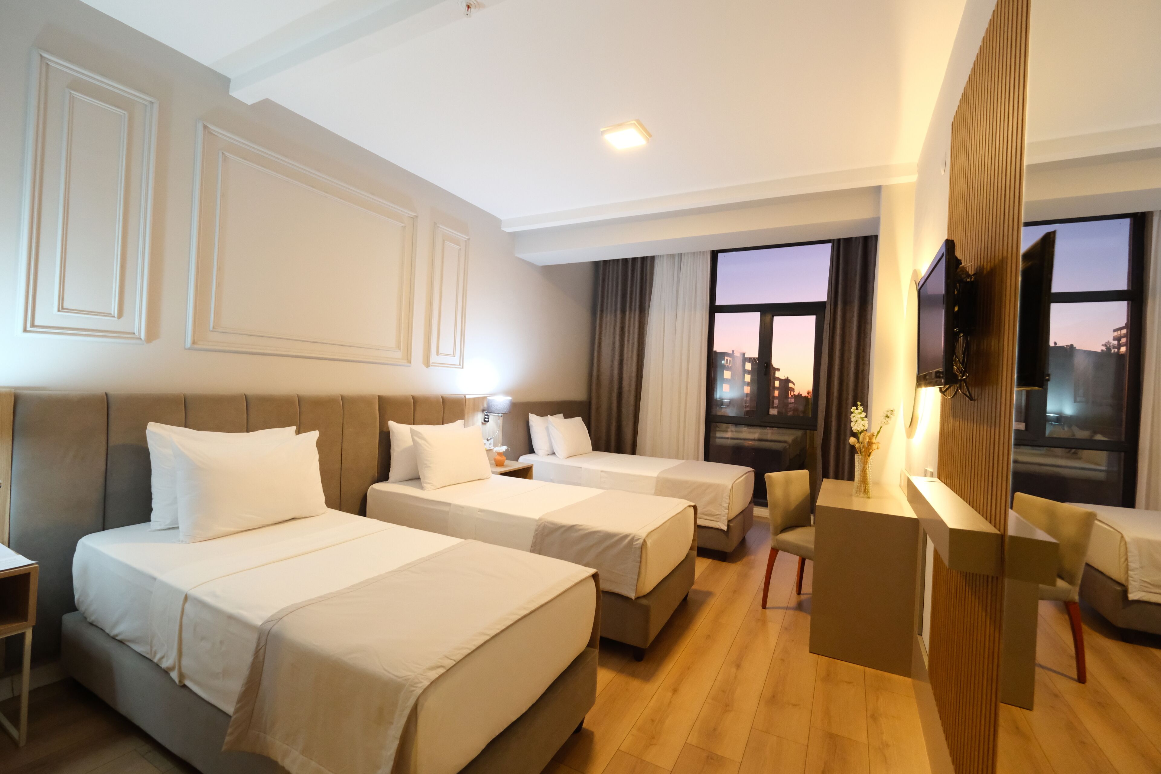 A hotel room with two beds, a desk, a TV, and a view of the city at sunset.