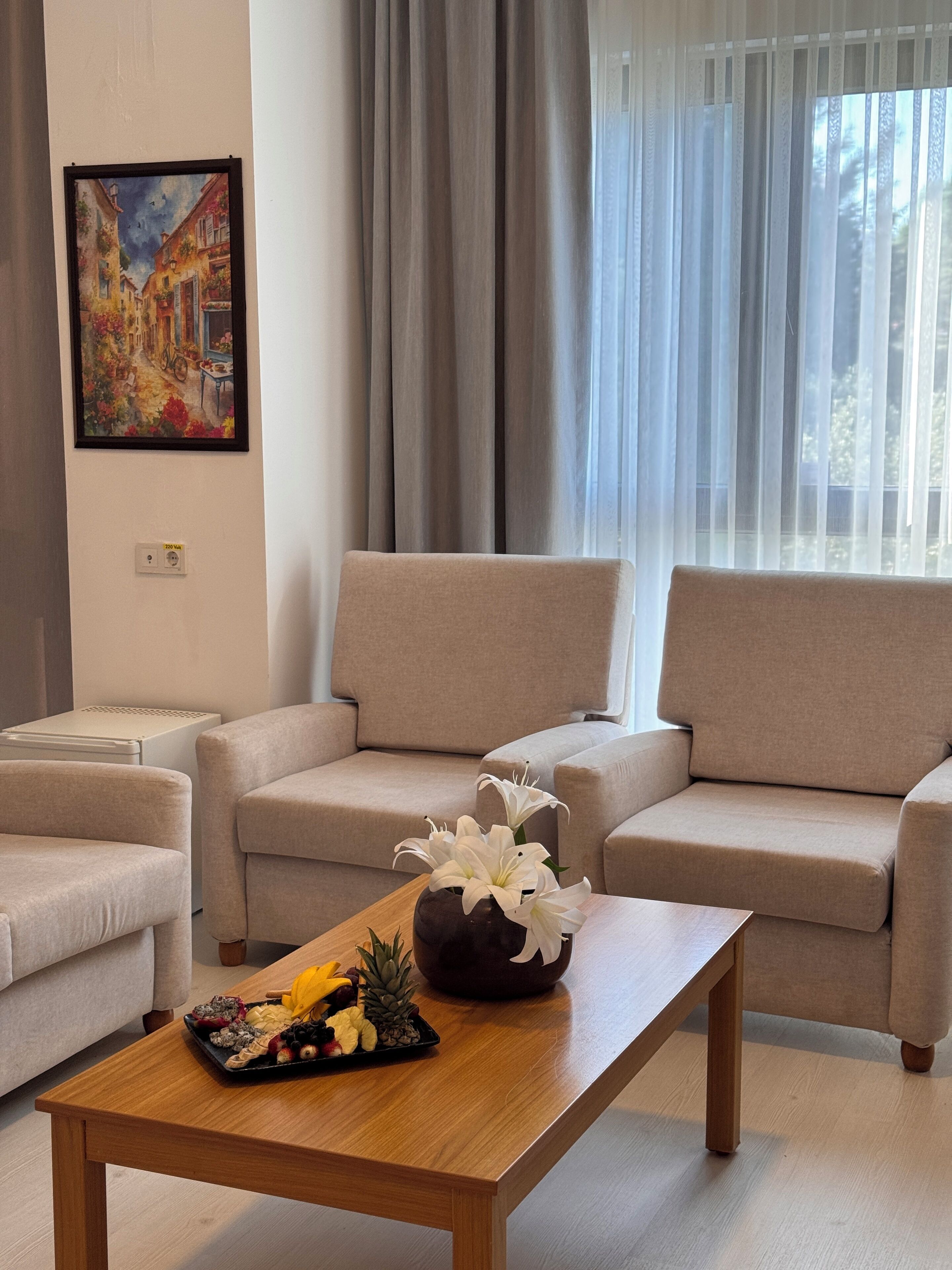 Un salón con un sofá beige, una mesa de centro de madera con fruta y flores, y un cuadro en la pared.