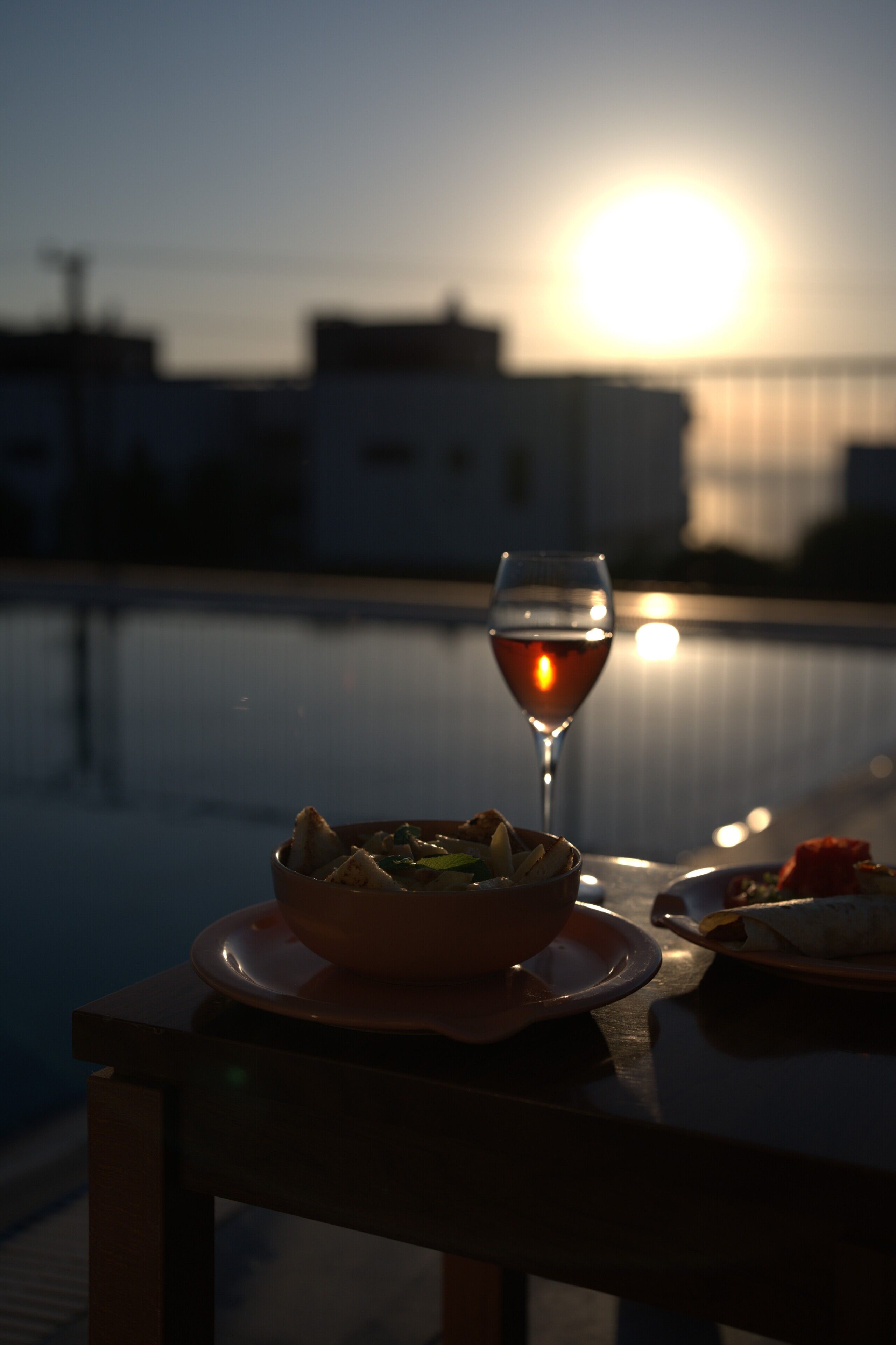A dining table with a bowl of food and a glass of red wine, set against a backdrop of a sunset over a body of water.