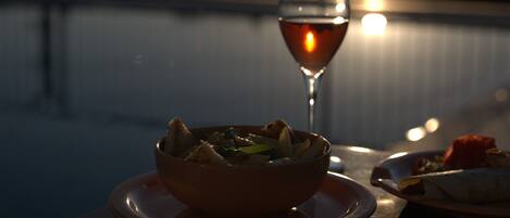 A dining table with a bowl of food and a glass of red wine, set against a backdrop of a sunset over a body of water.