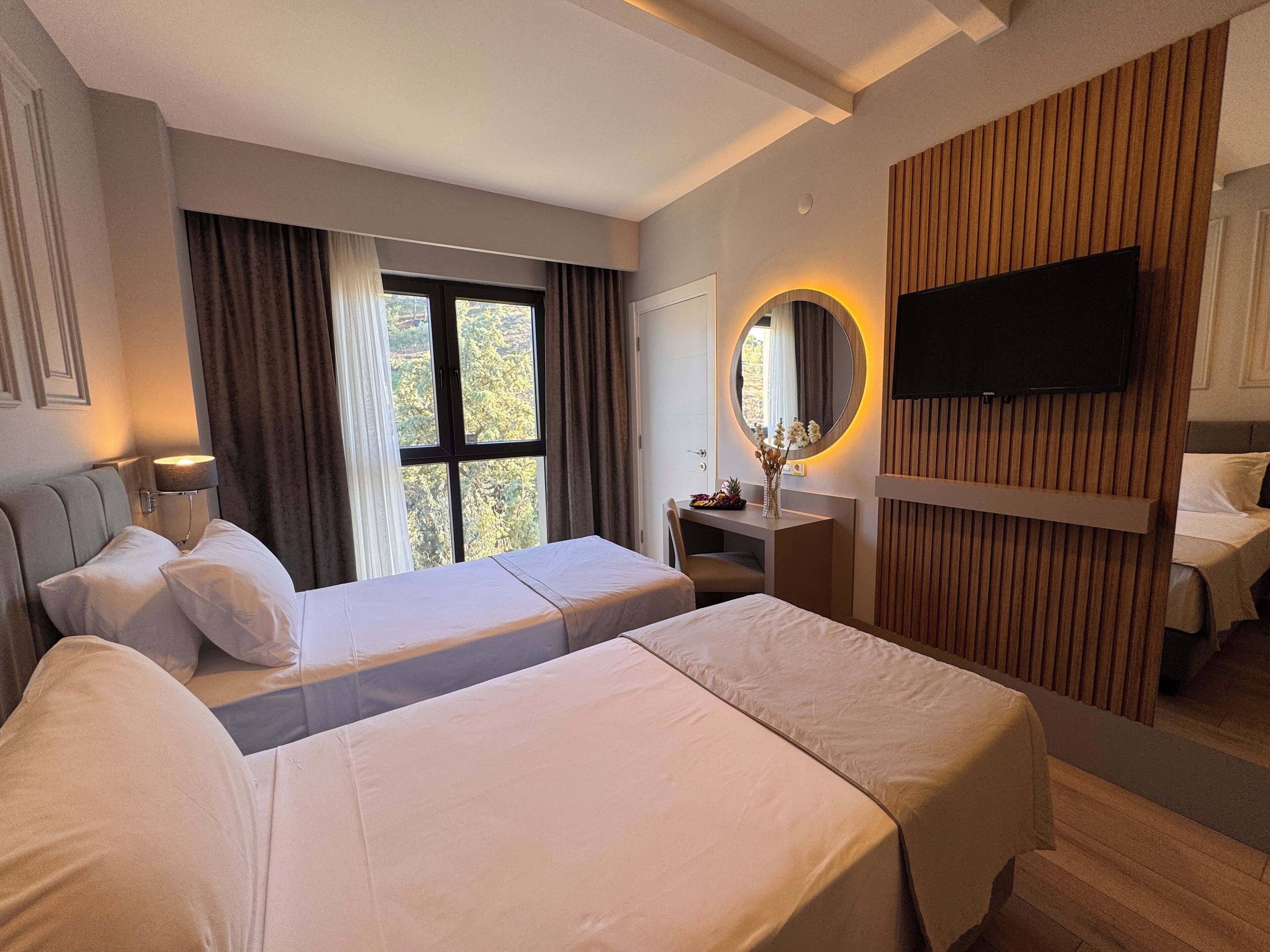 A hotel room with two beds, a desk, a TV, and a large window with curtains.
