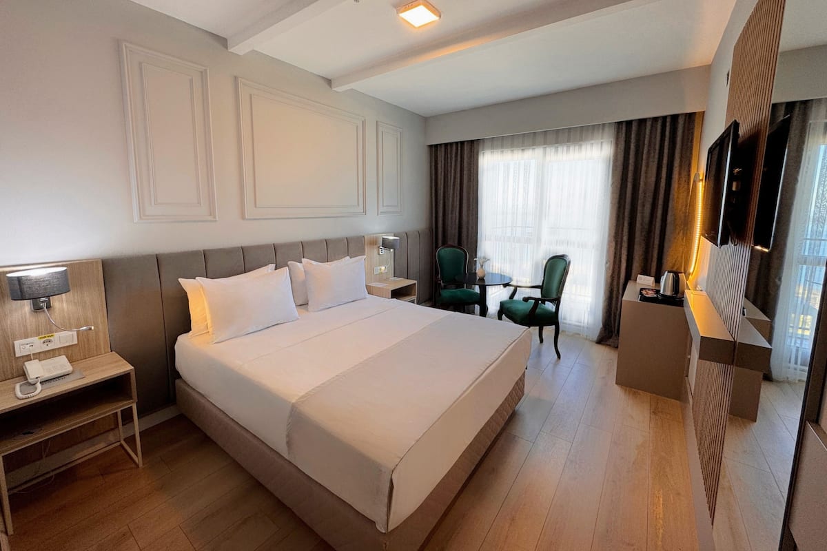 A hotel room with a large bed, a bedside table with a lamp, a small table with two chairs, a TV, and a window with curtains.