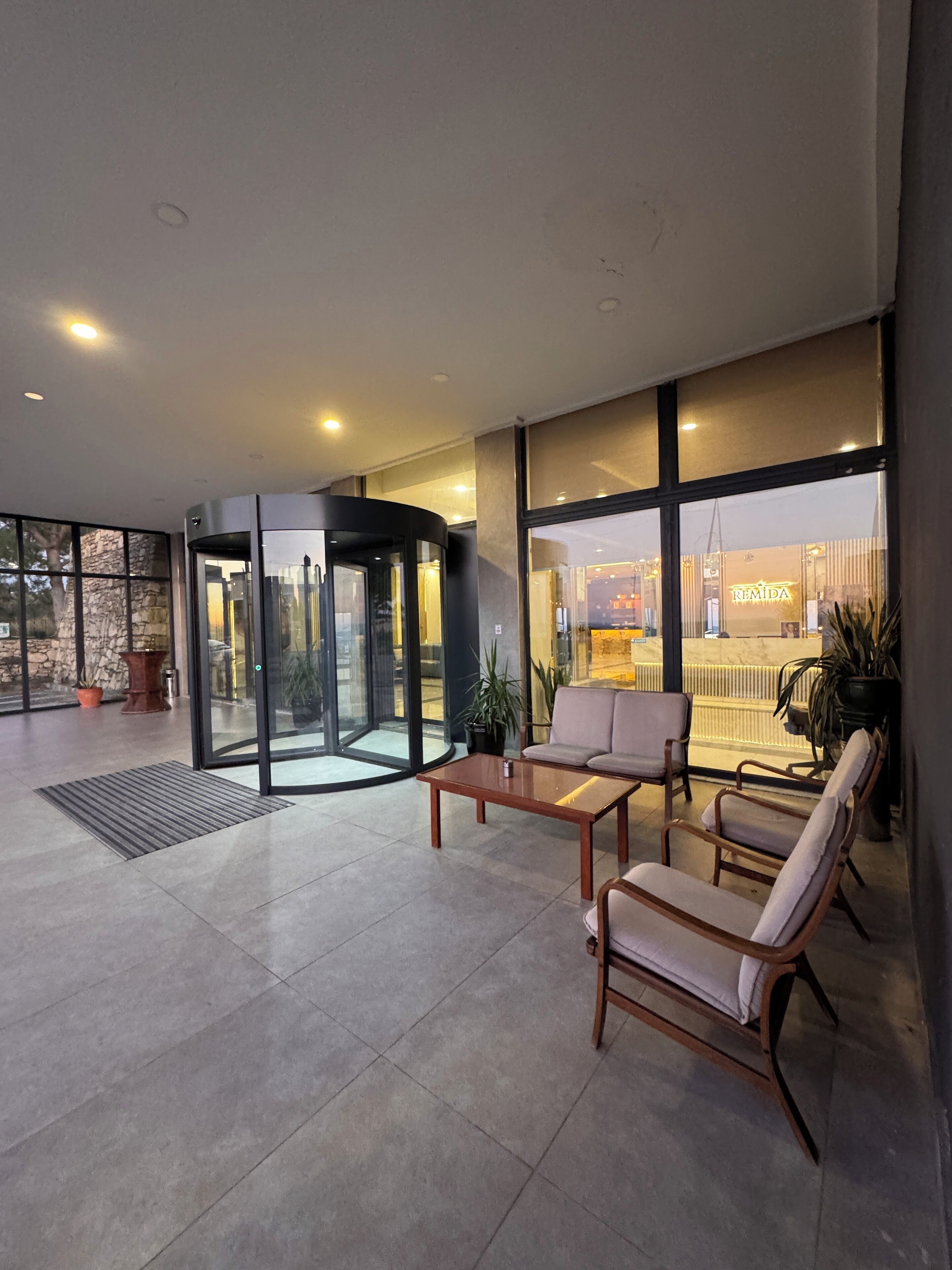 Un vestíbulo moderno con puerta giratoria, zona de estar y grandes ventanales que ofrecen vistas al exterior.