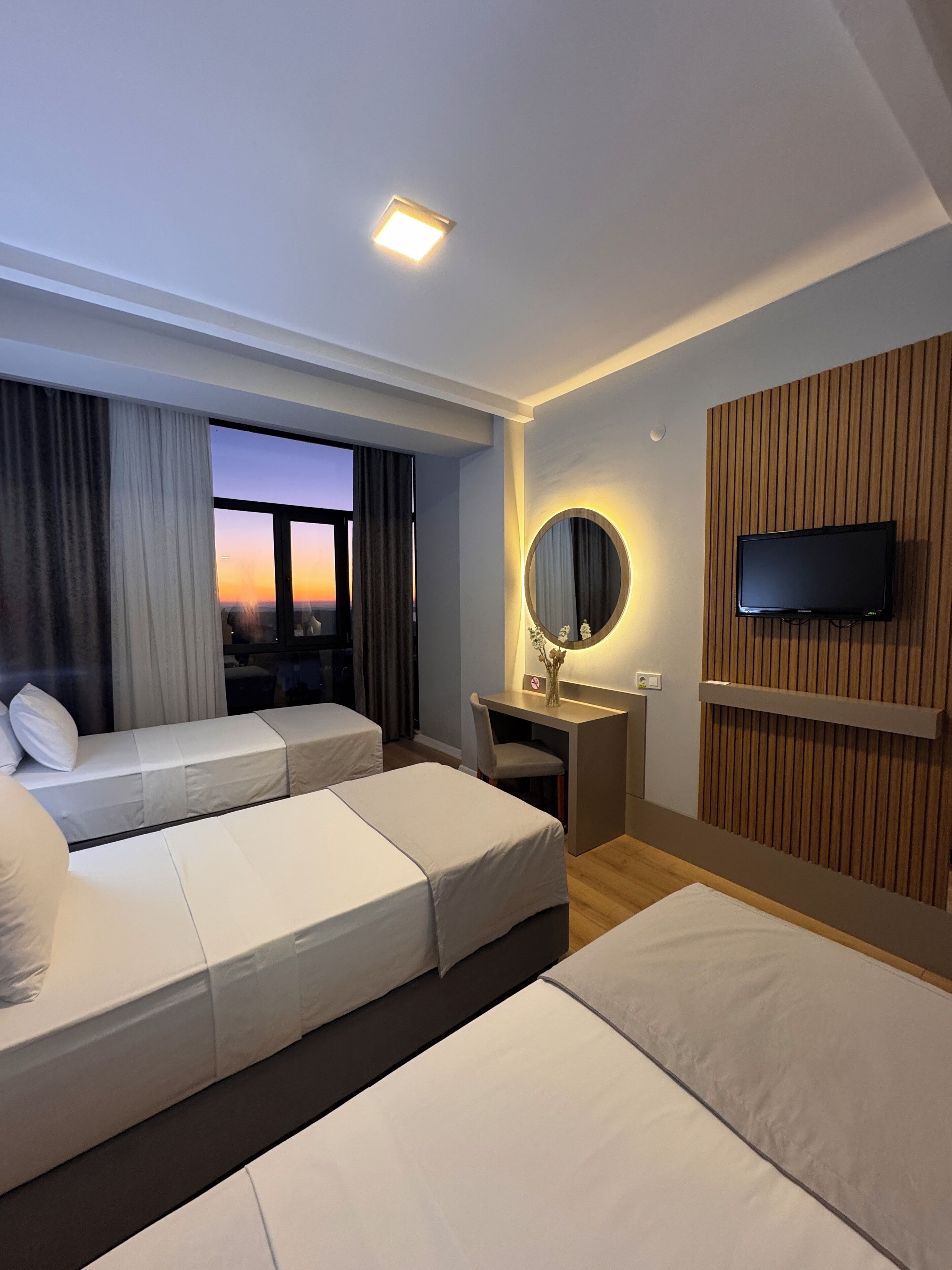 A hotel room with two single beds, a desk with a chair, a TV mounted on a wooden panel wall, a large window with curtains, and a ceiling light.