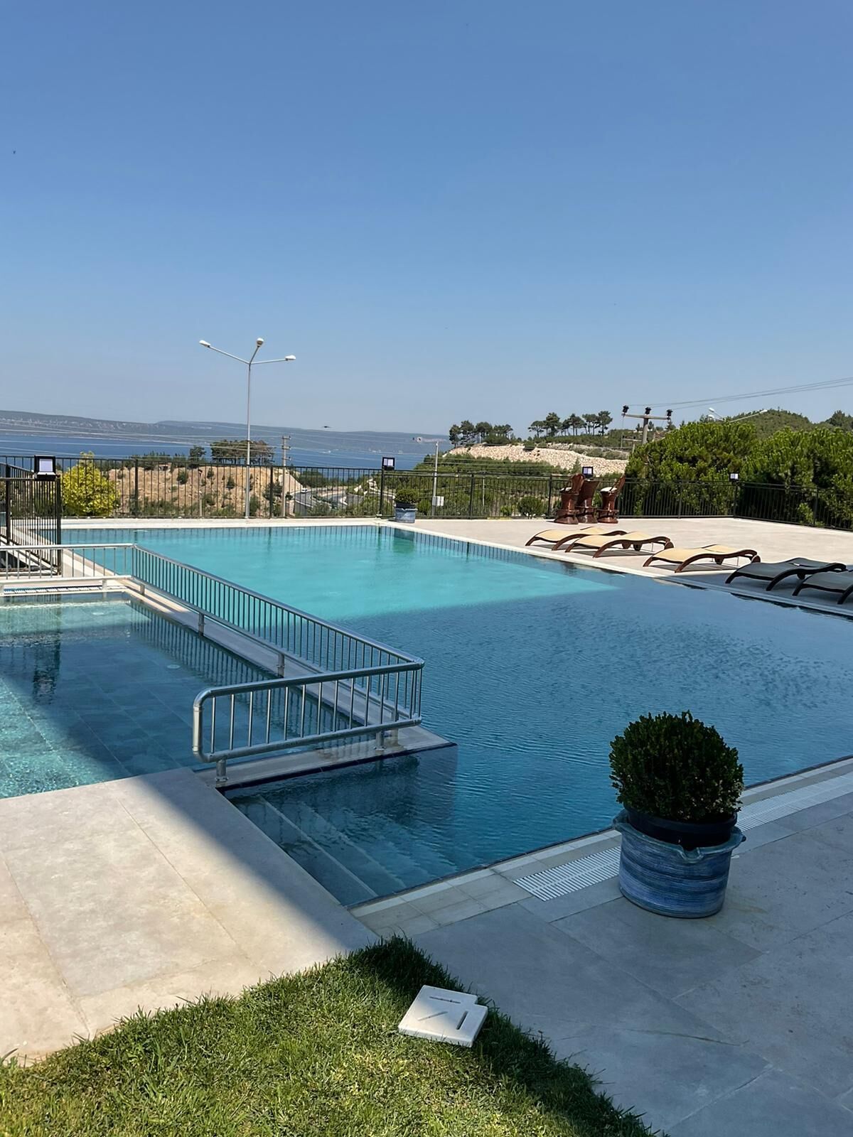 A rectangular swimming pool with a metal railing, a small grassy area with a potted plant, and a view of the sea and distant landscape.