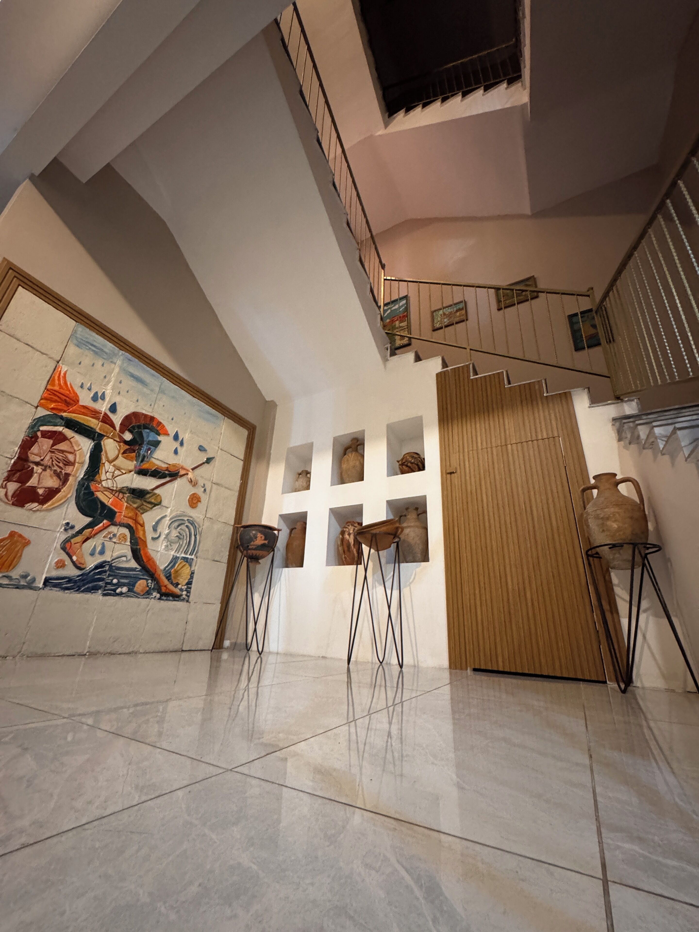 Un pasillo amplio con suelo de baldosas, un mural grande y una escalera con barandilla.