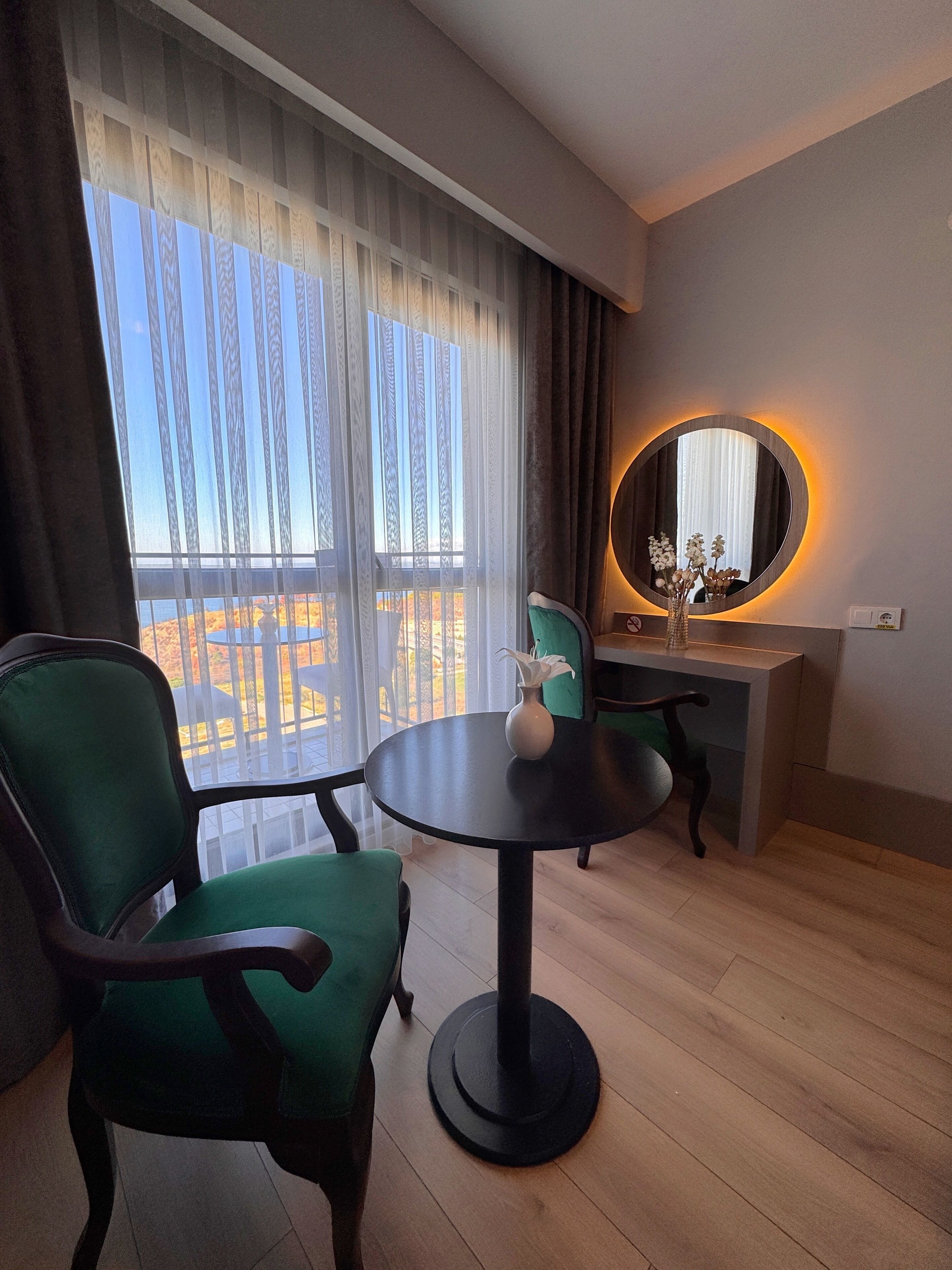 Une chambre d’hôtel avec une table ronde, deux chaises vertes, un bureau avec une chaise, un miroir rond avec un éclairage et une vue sur un balcon doté d’une balustrade.