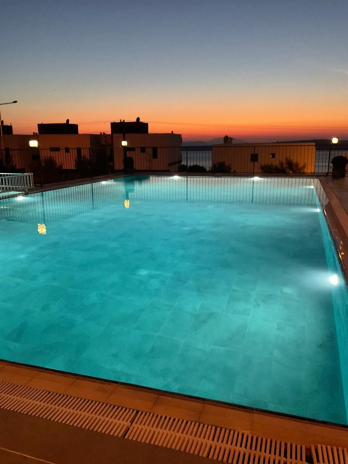 A rectangular swimming pool with clear blue water, surrounded by a low fence and a series of buildings in the background.