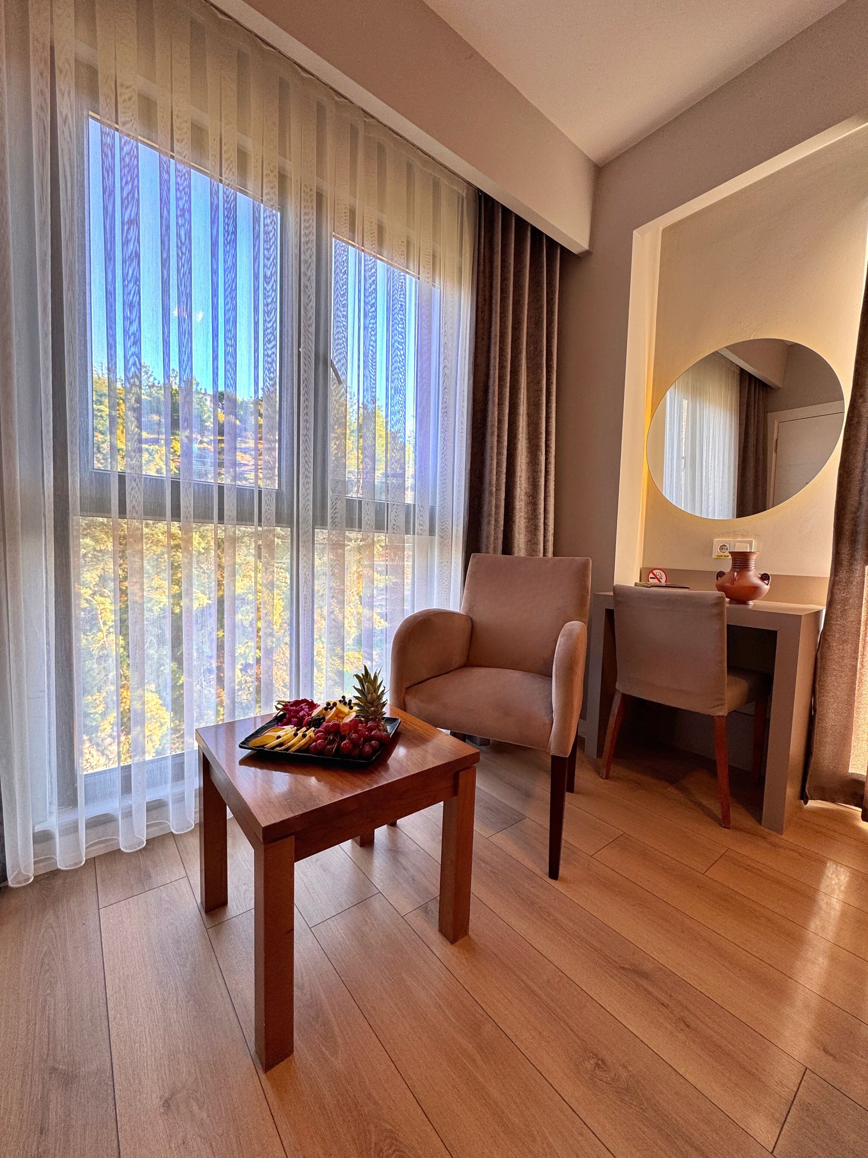 Habitación de hotel con piso de madera, una mesa de comedor con fruta, una silla, un escritorio con espejo y ventanas grandes con cortinas traslúcidas.