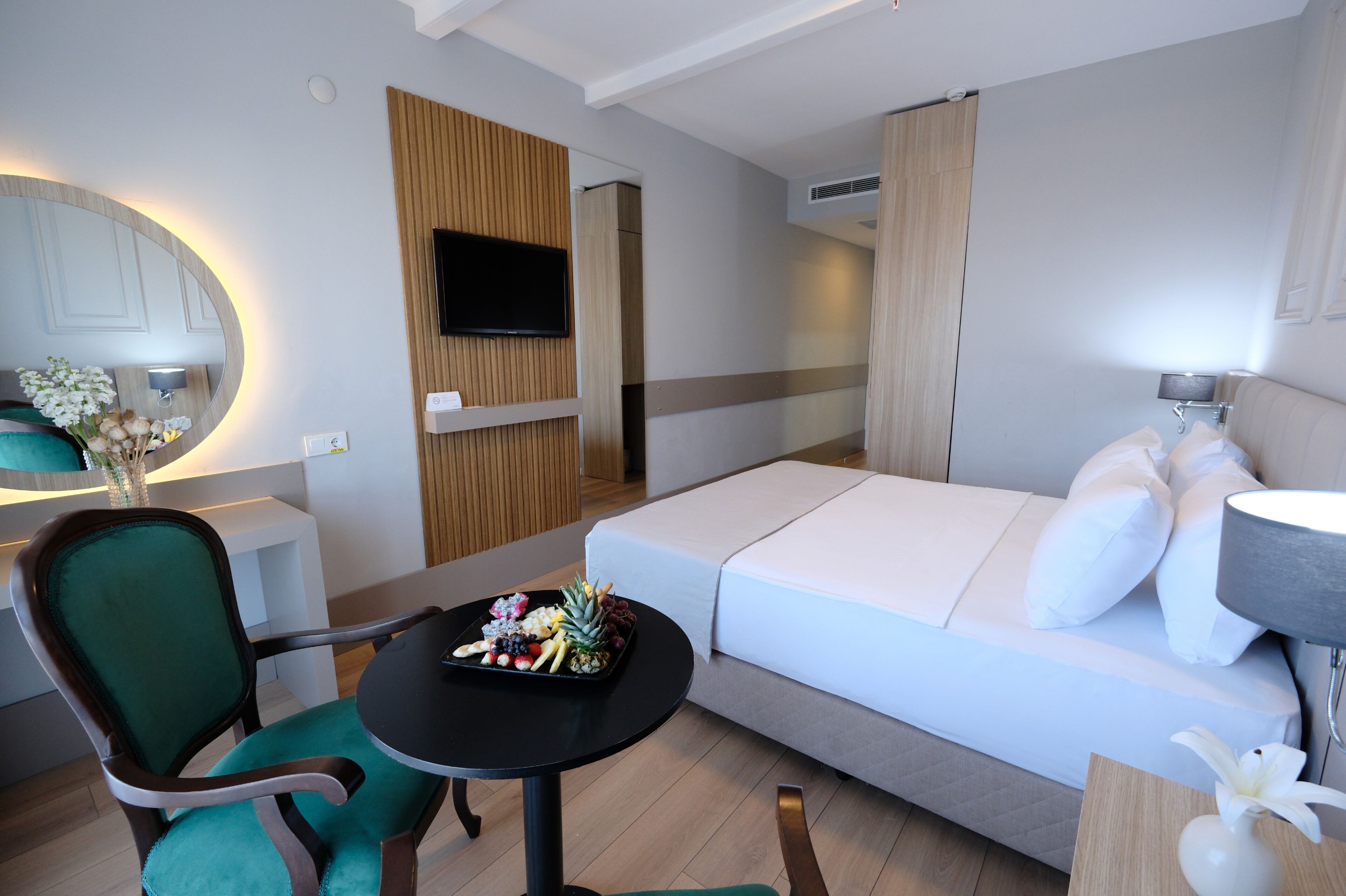 Et moderne hotelværelse med en seng, et lille bord med et frugtfad, en grøn stol, et fjernsyn ophængt på væggen og et spejl.