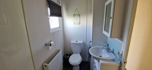 A small bathroom with a toilet, a sink, a radiator, and a window with a blind. - Beachcomber Cabin Dog Friendly - Anderby Creek (Skegness)