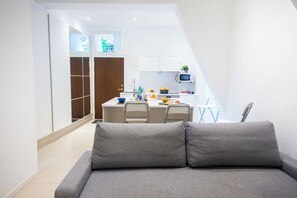 Un apartamento moderno con un sofá gris en primer plano, una cocina con armarios blancos y un microondas, y un guardarropa de madera.