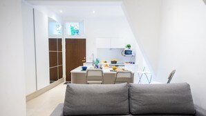 Een modern appartement met een grijze bankstel op de voorgrond, een keuken met witte kasten en een magnetron, en een houten garderobe.