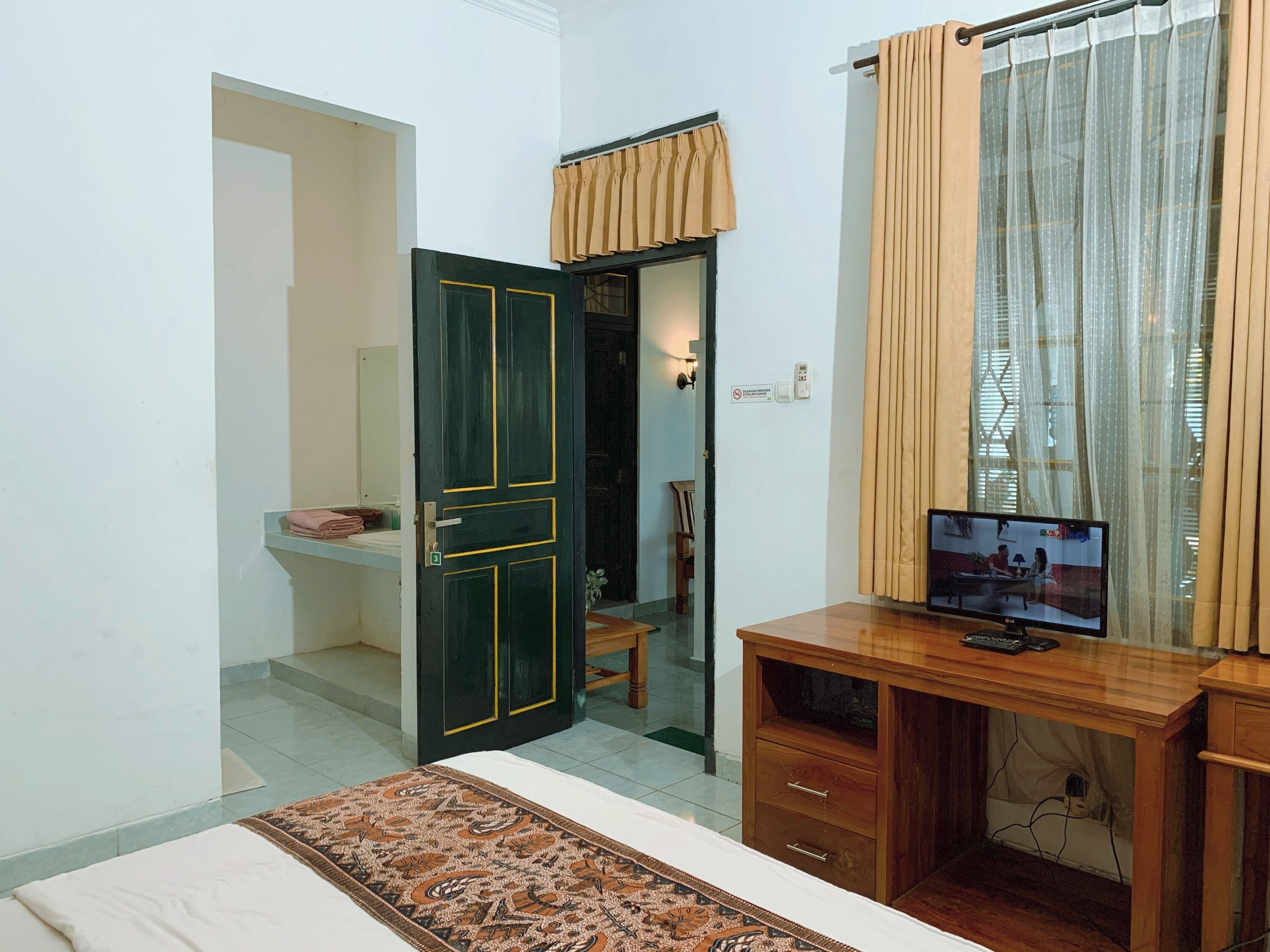 A bedroom with a bed, a desk with a computer, a mirror, and a door leading to another room.