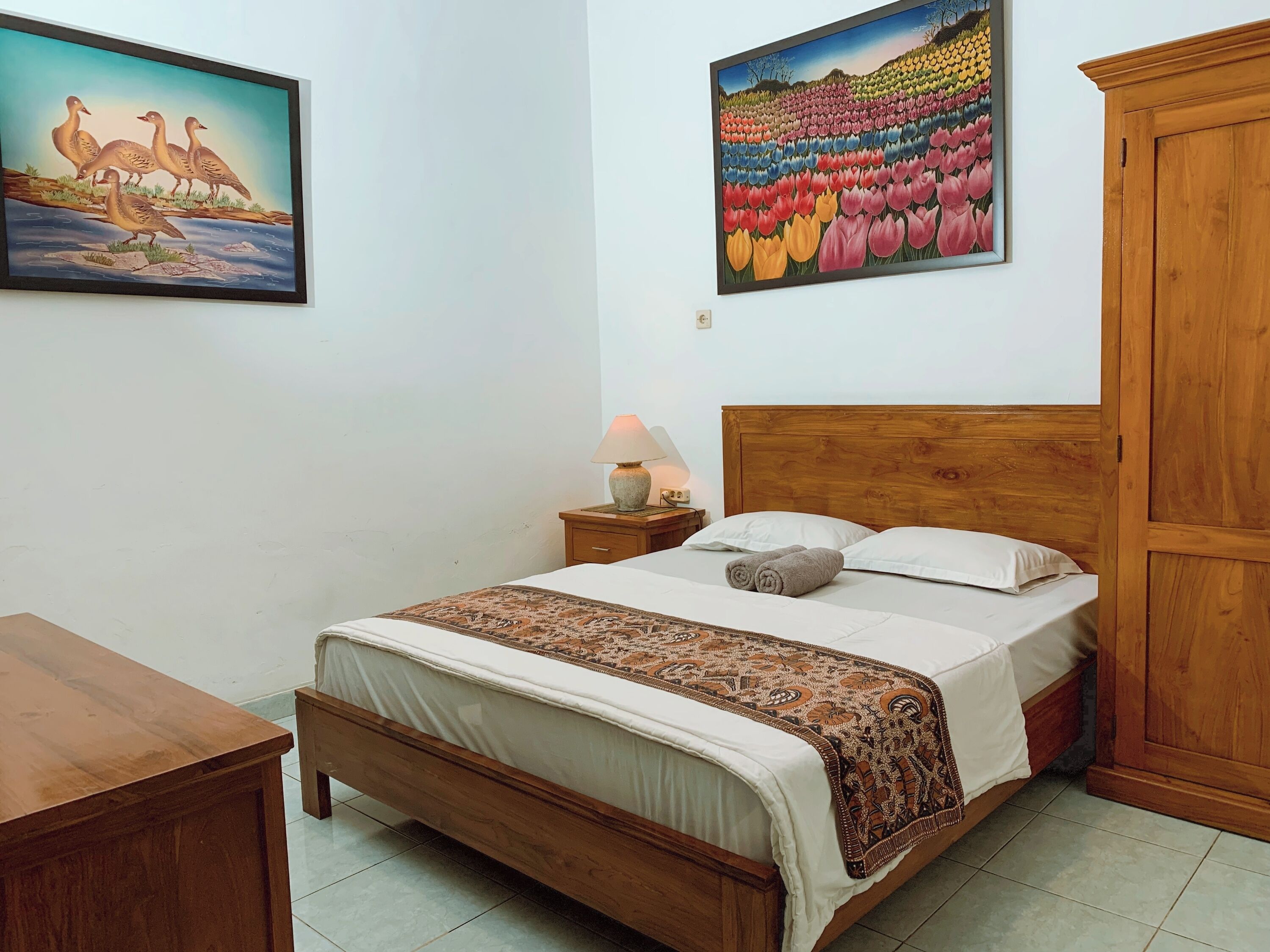 A bedroom with a wooden bed, a nightstand with a lamp, a wooden wardrobe, and two paintings on the wall.