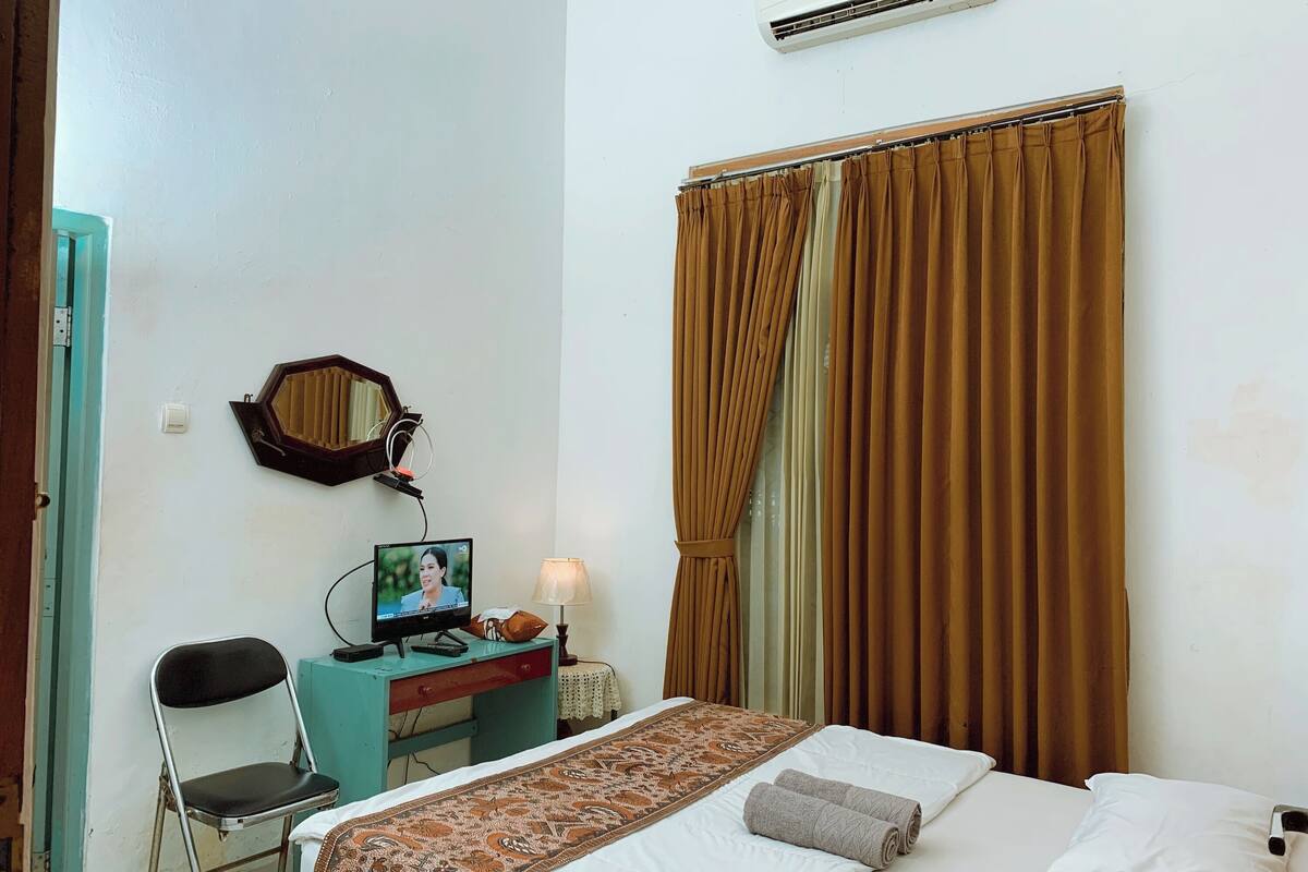 Una habitació d'hotel amb un llit, una taula de treball amb un ordinador, una cadira, una làmpada i una finestra amb cortines.