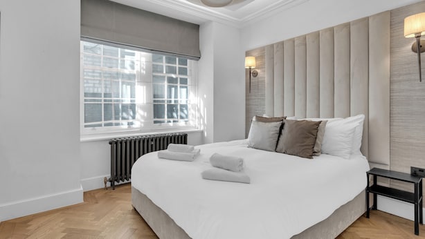 A modern bedroom with a large bed, a radiator, a window with blinds, and a wall-mounted light fixture.