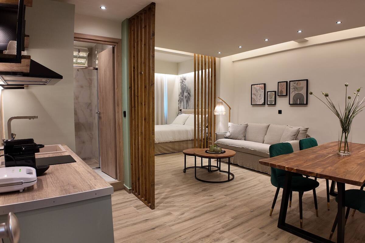 A modern kitchen with a built-in stove, a dining area with a wooden table and green chairs, and a bedroom with a bed and a window.