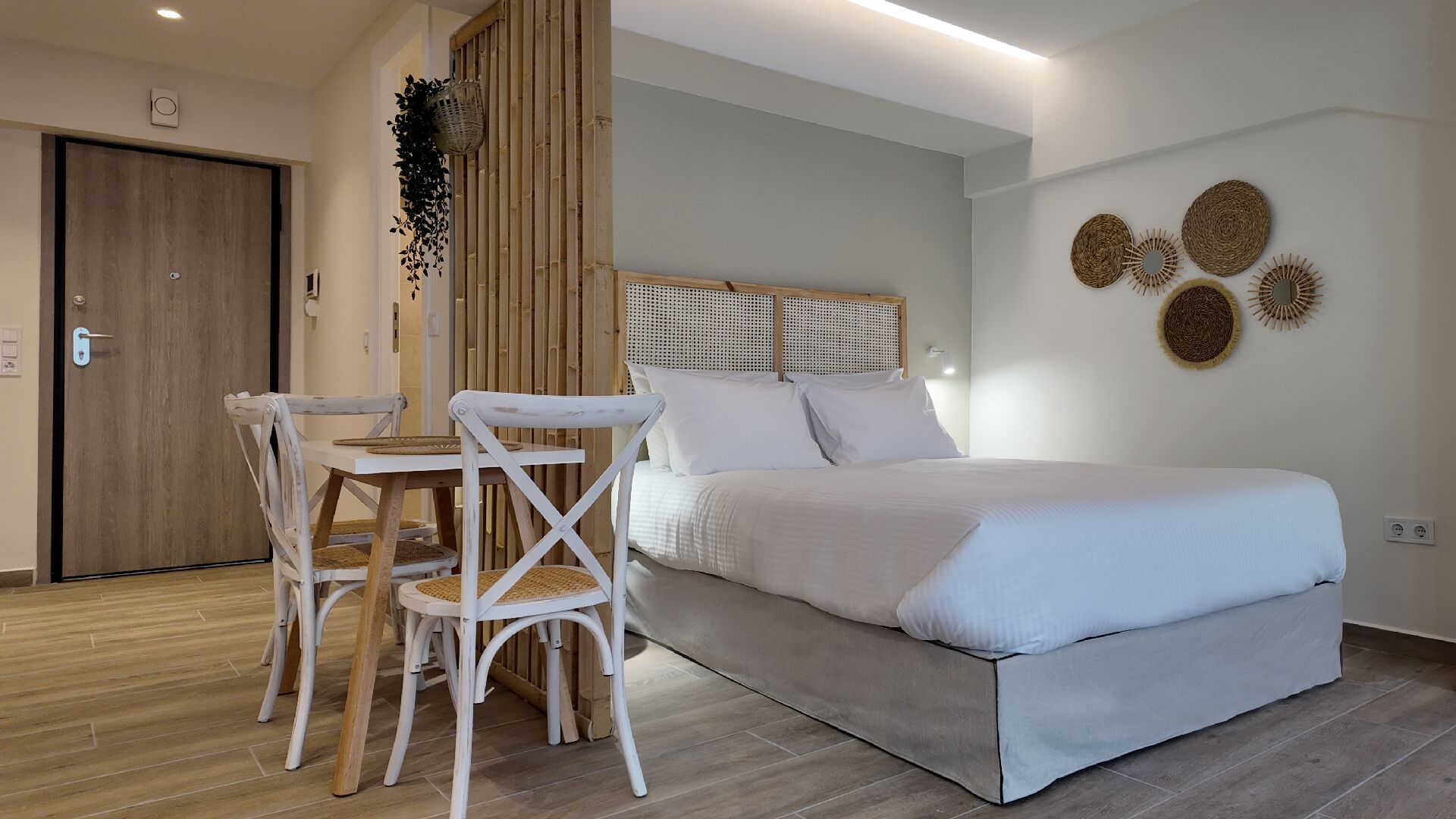 A bedroom with a bed, a wooden door, a dining table with chairs, and a wall with decorative items.