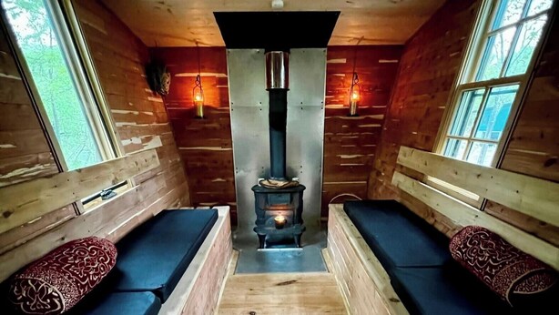 A cabin with a central wood-burning stove, two benches with patterned cushions, and large windows.