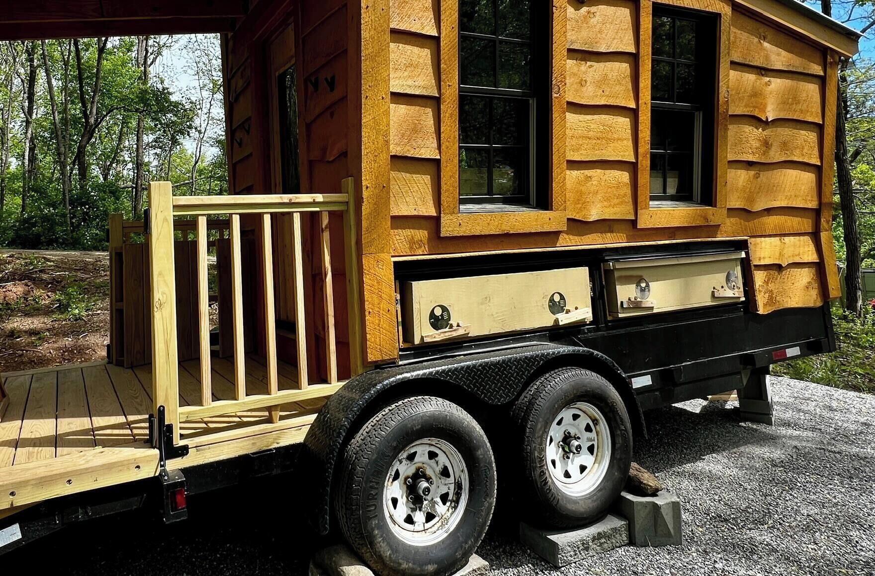 A wooden mobile home with a flatbed trailer, windows, and a small deck.