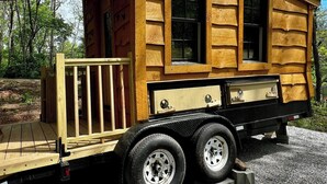 A wooden mobile home with a flatbed trailer, windows, and a small deck.