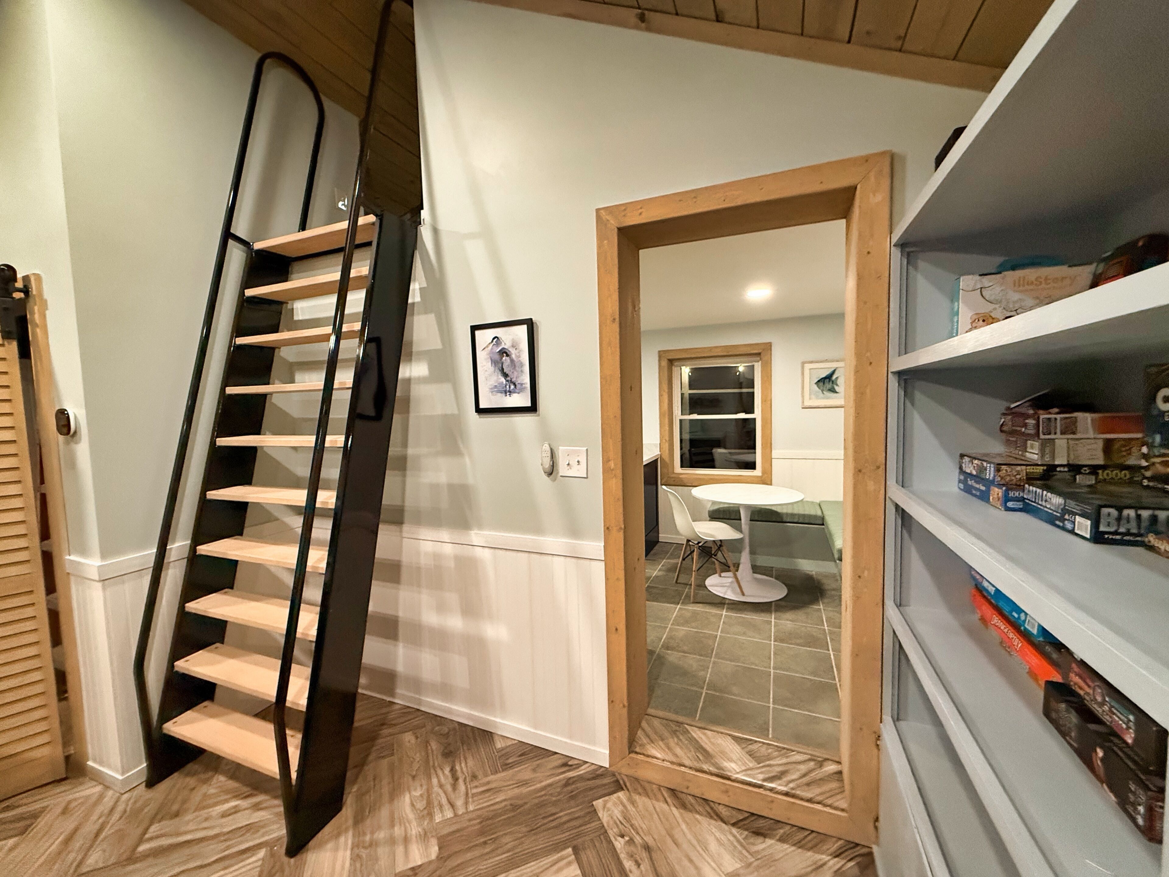 A staircase with a wooden finish, a dining area with a white table and chairs, and a shelf with board games.