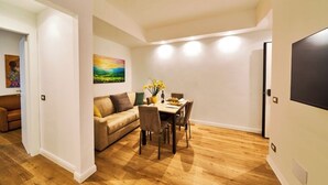A modern living room with a wooden floor, a beige sofa, a dining table with chairs, and a flat-screen TV mounted on the wall.