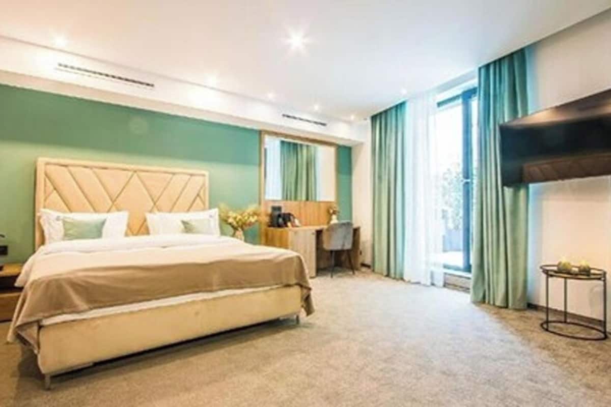 A hotel room with a bed, bedside table, desk, and a large window with curtains.