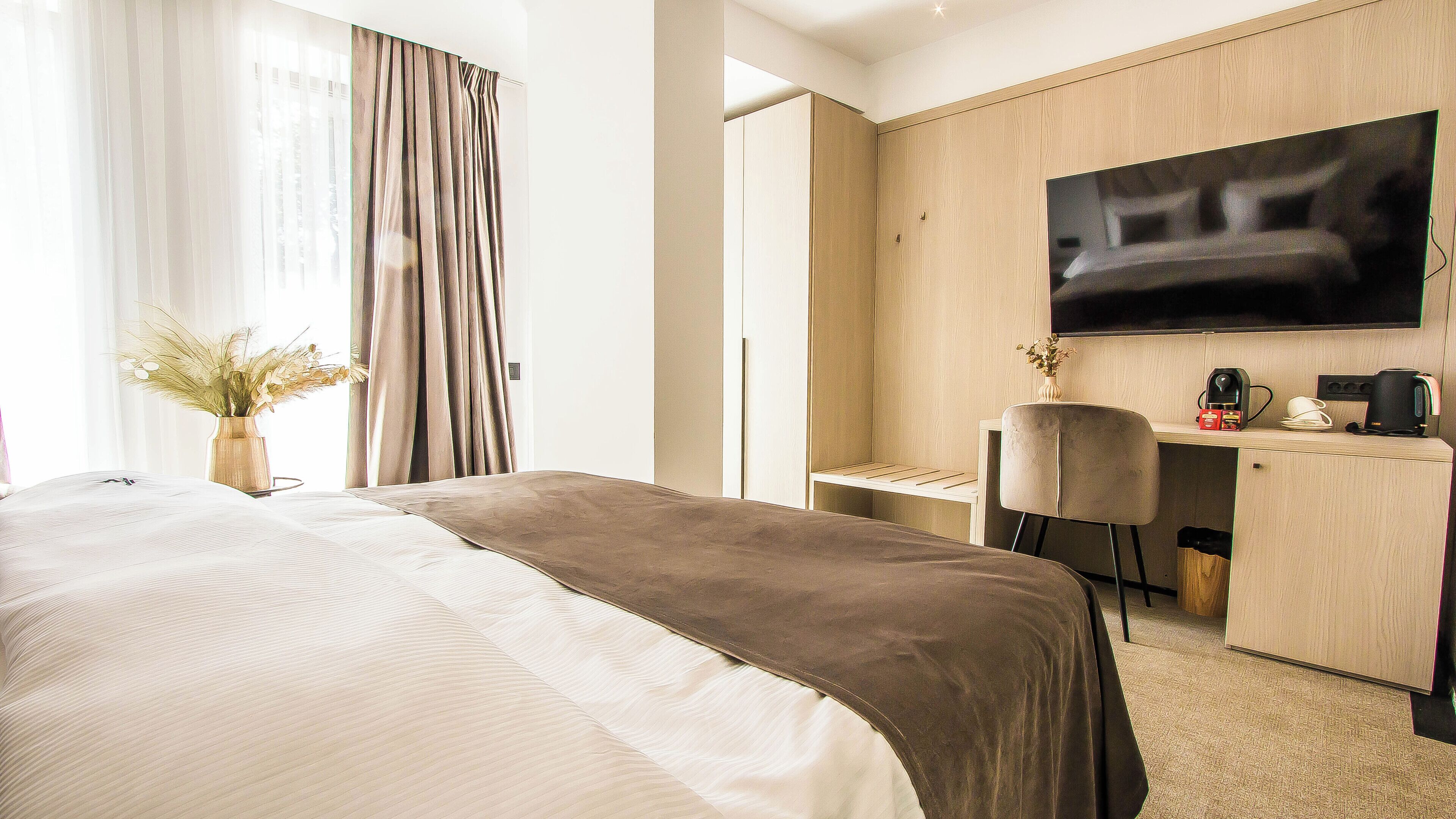 A hotel room with a bed, a desk, a chair, a flat-screen TV, and a vase with dried flowers.