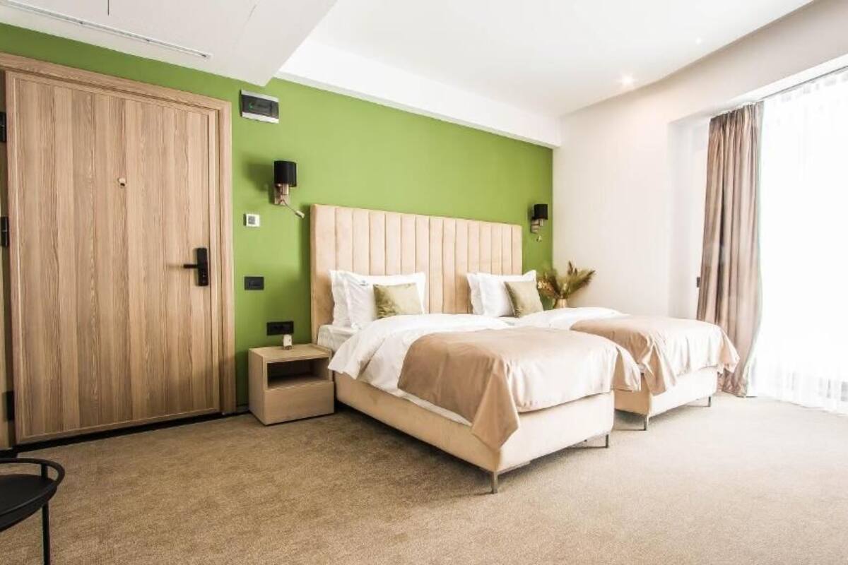 A hotel room with two beds, a wardrobe, bedside tables, and a large window with curtains.