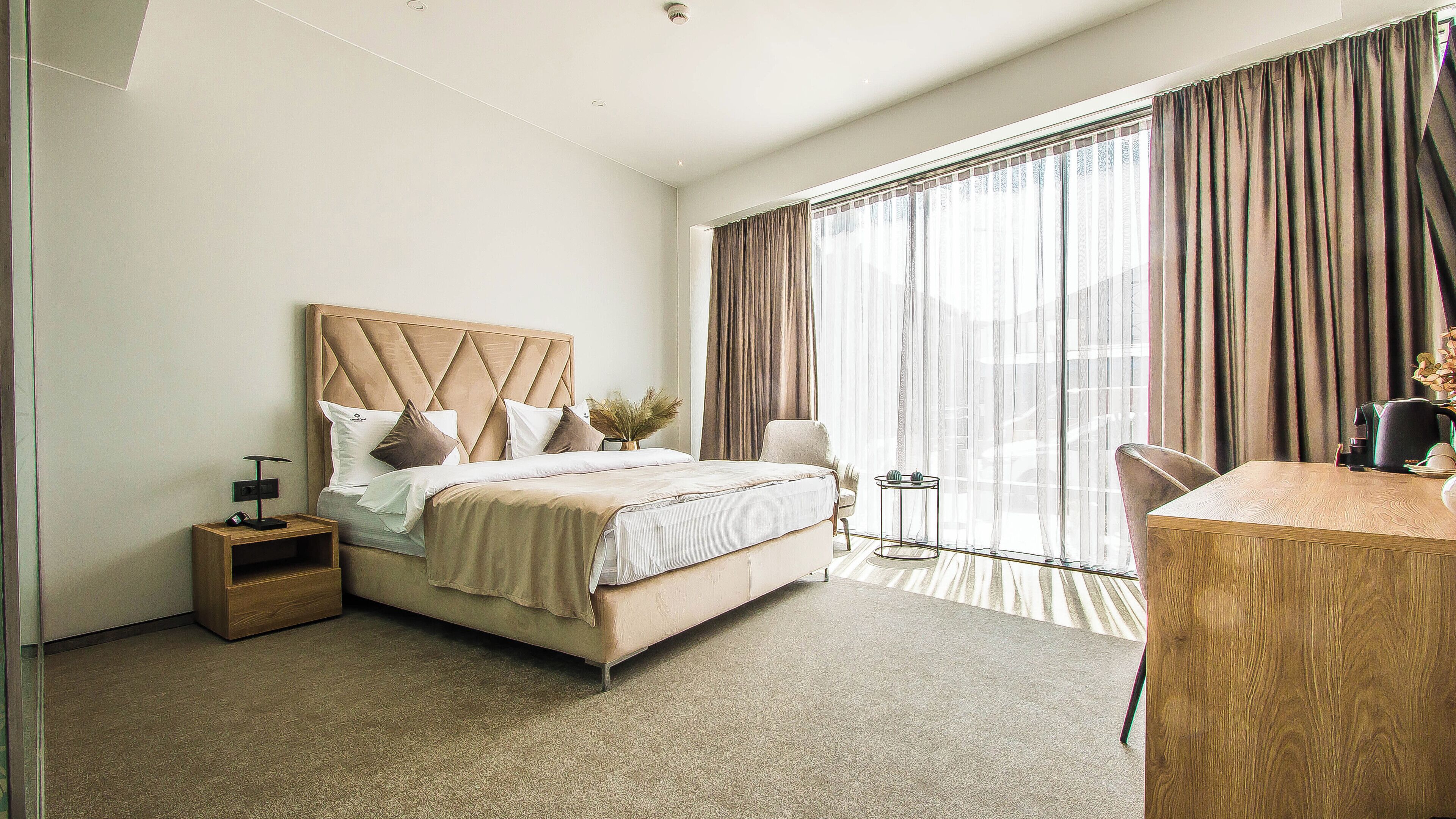 A modern hotel room with a large bed, a wooden desk, and a view of the city through sheer curtains.