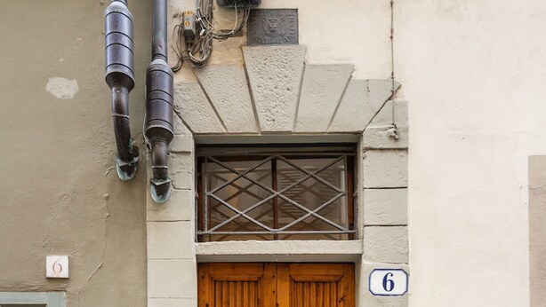 Eine Holztür mit Oberlicht, einem Schild mit der Nummer 6 und einem Gulideckel auf dem Boden.