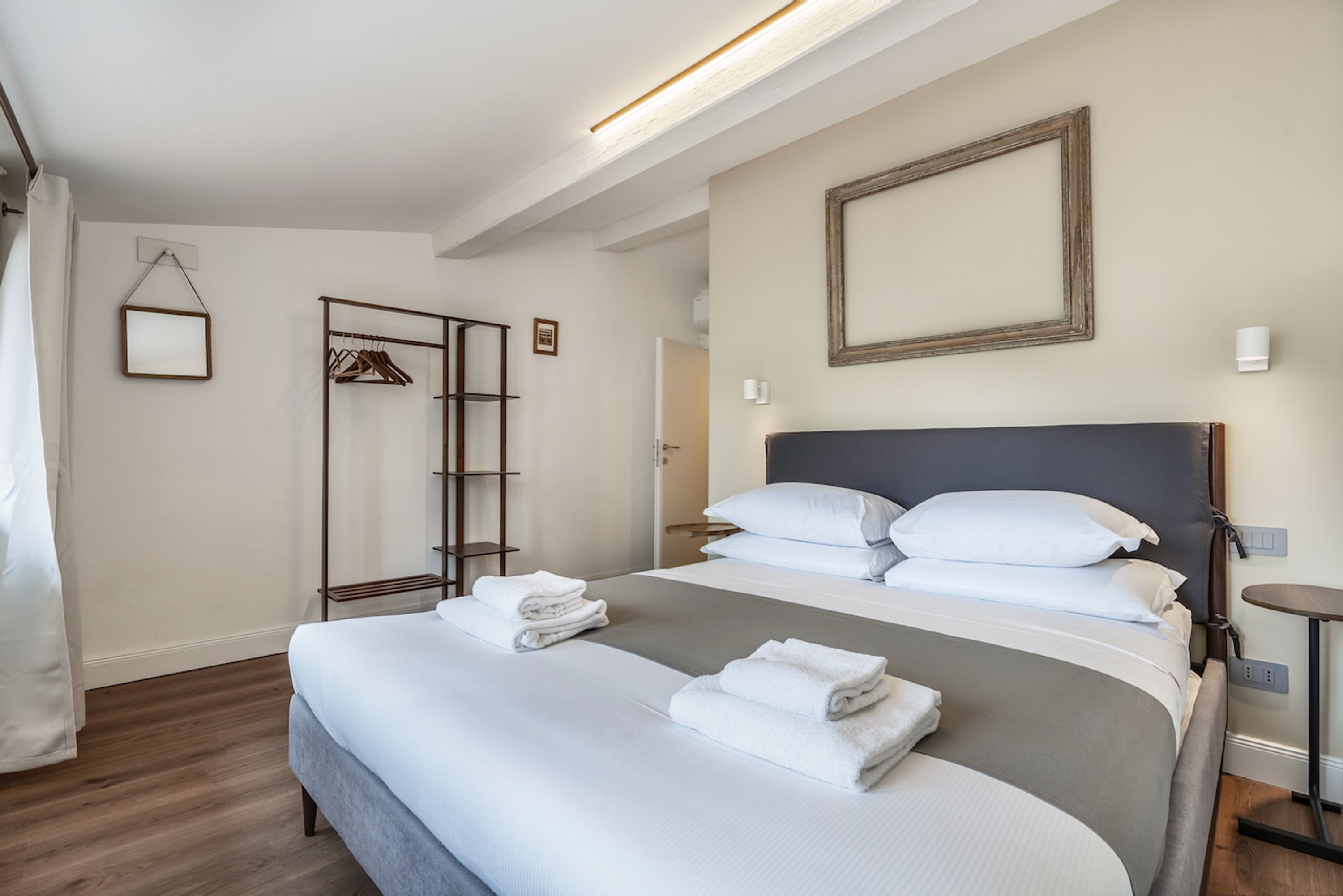A neatly made bed with white linens and towels, a wooden floor, a coat rack, and a framed picture on the wall.