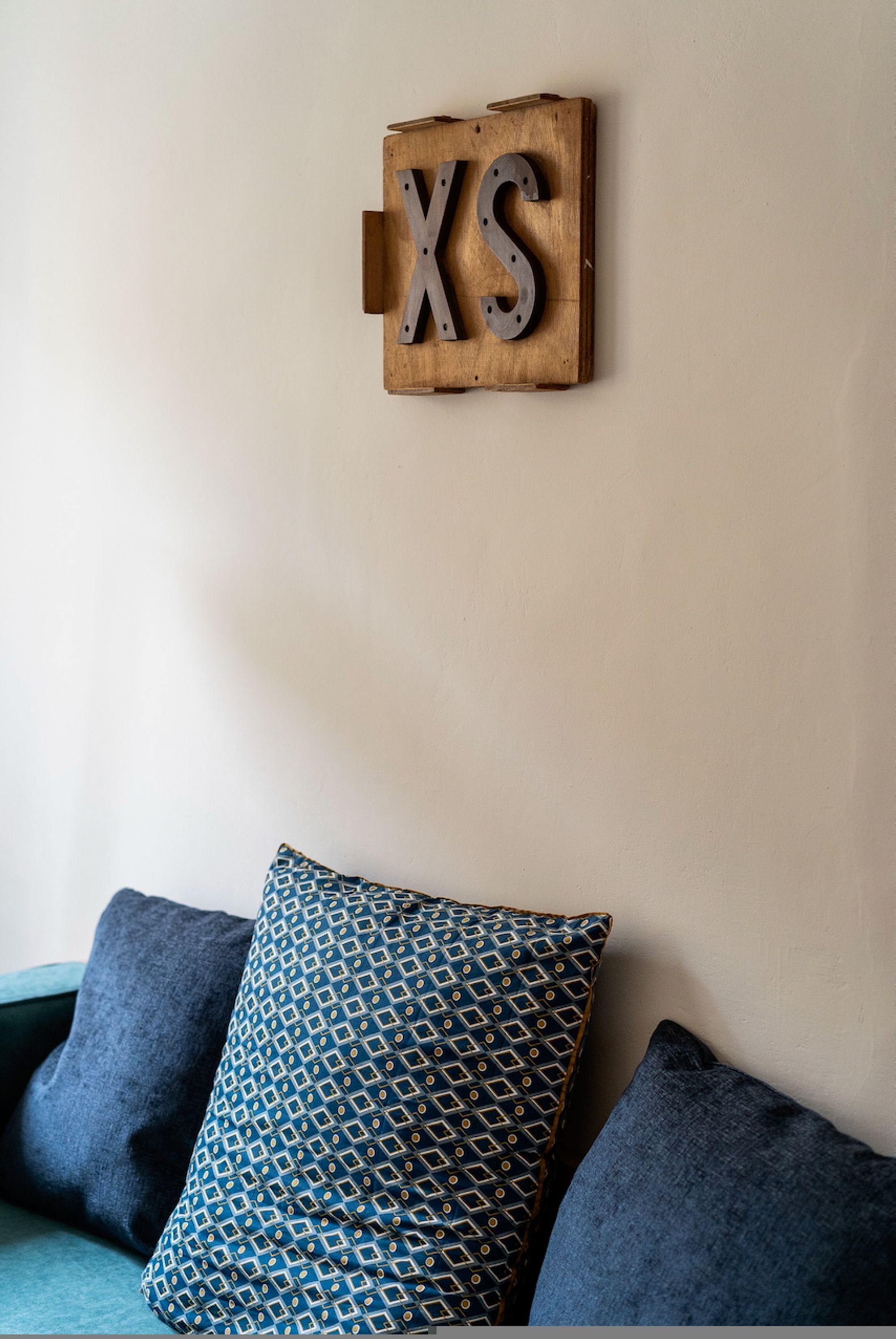 A wooden sign with the letters "XS" mounted on a wall above a sofa with patterned cushions.
